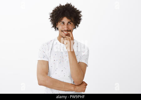 Man laughing over his own thought while thinking, smiling joyfully, holding finger on lip and gazing at upper right corner, imaging funny moment happened with boss, standing pleased over grey wall Stock Photo