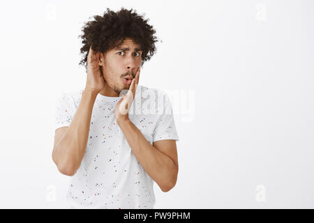 Portrait of gossiping stunned man. Portrait of shocked and stunned emotive curly-haired hispanic guy, frowning from shook, covering mouth with palms while hearing disgusting rumor Stock Photo