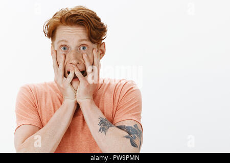 Portrait of redhead young man with freckles covering face with palms and peeking through fingers, screaming from fear and staring frightened at camera, scared of horror movies, posing over gray wall Stock Photo