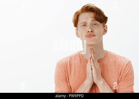 Please lend me hand. Portrait of cute young caucasian male with freckles and red hair, pursing and folding lips, lifting chin up and holding hands in pray while begging for help or favor Stock Photo
