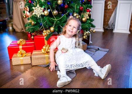 cute little girl in a white dress sits near a Christmas tree with gifts and looks up Stock Photo