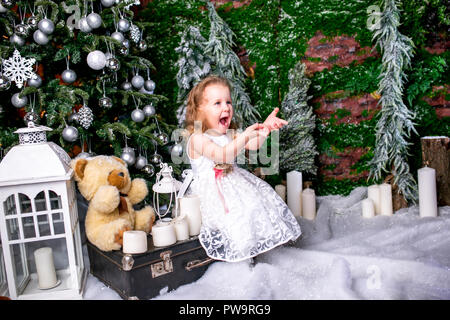 cute little girl in a white dress sitting near a Christmas tree on a suitcase next to the candles and a teddy bear, throws snow up and laughs Stock Photo