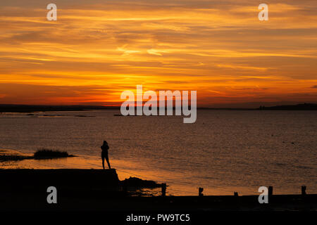 A woman stands on a breakwater, admiring the sunset at Seasalter, Whitstable, Kent, UK. She is looking across the Swale estuary towards the Isle of Sheppey. Stock Photo