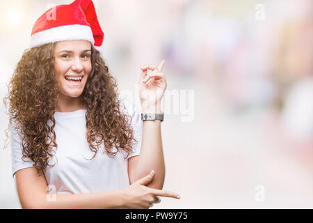 Young brunette girl wearing christmas hat over isolated background with a big smile on face, pointing with hand and finger to the side looking at the  Stock Photo