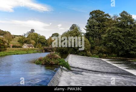 Water from the River Derwent rushes down a weir towards a woodland.   A sluice gate and small building are on the far bank. Stock Photo