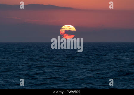 A new day as a bright orange sun rises above the sea on the horizon with wispy clouds in front set against a pink and orange sky at dawn Stock Photo