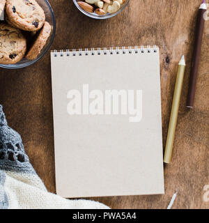 Flat lay of blank brown notebook with knitted blanket and snacks on wooden table. Cozy autumn or winter mood. Copy space. Stock Photo