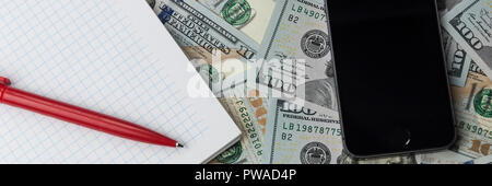 A telephone, a notebook and a pen lay on scattered hundred dollar bills. Stock Photo