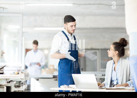 Young engineer in gloves and overalls talking to his colleague sitting by desk in front of laptop Stock Photo