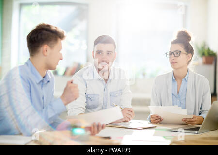Meeting of three young professionals with papers sitting by table and discussing information Stock Photo