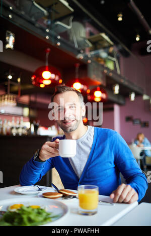 Happy young man with cup of drink sitting by served table in restaurant and having lunch Stock Photo