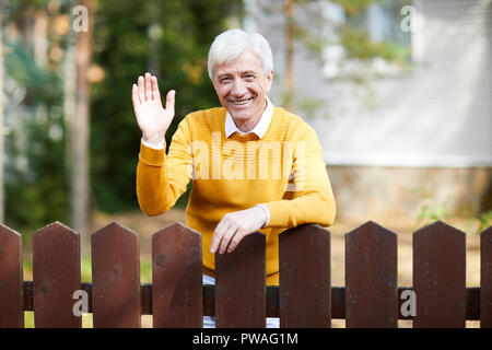 Cheerful senior man in casualwear standing by wooden fence and waving his hand to you Stock Photo
