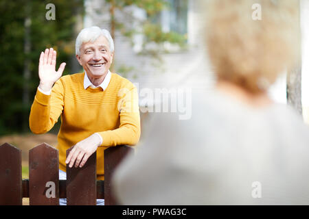Cheerful and friendly mature man waving hand to his neighbour while standing by fence Stock Photo