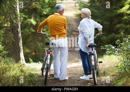 Rear view of senior active couple with bicycles having talk while moving along forest path Stock Photo