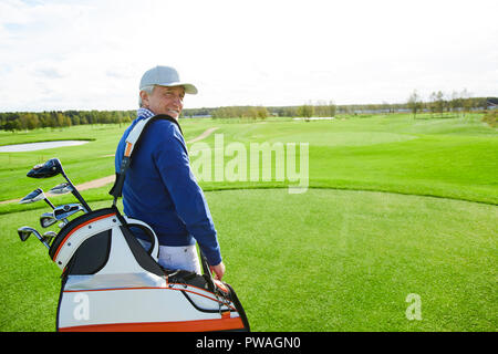 Happy senior man with bunch of golf clubs in bag looking at camera while walking down green field Stock Photo