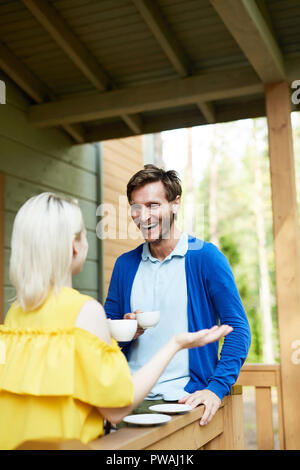 Excited young man with cup of drink laughing during talk to his wife on terrace of country house Stock Photo