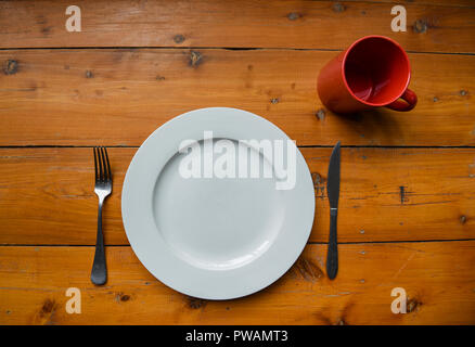 Top view of clean round white plate, silver fork and knife, red mug on a wooden table Stock Photo