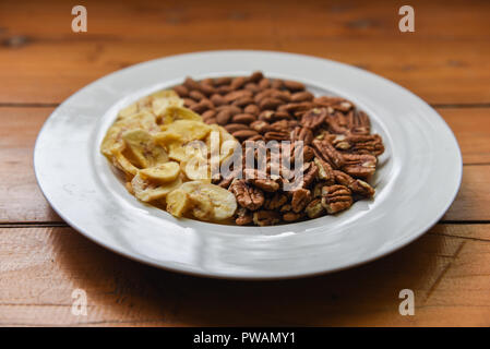 Healthy dried bananas, pecan nuts and almond nuts in the middle of a white plate on wooden table Stock Photo
