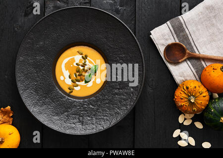 Pumpkin and carrot soup with pumpkin seeds, cream and basil in black modern plate on dark wooden background. Top view. Stock Photo