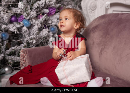 A beautiful cute little girl dressed in an elegant evening red dress sits on the couch and opens a New Year's gift. Against the background is a Christ Stock Photo