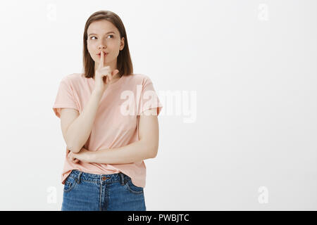 Girl shared secret asking not tell anyone else. Cute dreamy girlfriend in pink t-shirt, looking at upper right corner and smirking while making shush gesture with index finger over mouth, saying shh Stock Photo