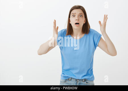 Portrait of anxious emotive young woman in blue t-shirt, shaking hands and gasping, explaining terrible news, being shocked and stunned with something horrible, standing over grey wall Stock Photo