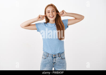Cannot hear what you say. Portrait of playful carefree and optimistic charming redhead female with freckles closing ears with index fingers and smiling from joy avoiding arguing over gray background Stock Photo