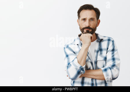 Hmm, doubt it. Portrait of serious handsome adult male teacher with beard in checked shirt, leaning head on fist and staring with strict expression at camera, listening carefully to explanation Stock Photo