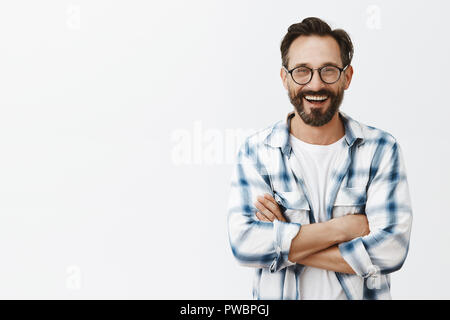 Charming male professor of university standing over gray background in stylish sunglasses and checked shirt, talking casually with colleagues about job and funny moments during tutoring, smiling Stock Photo