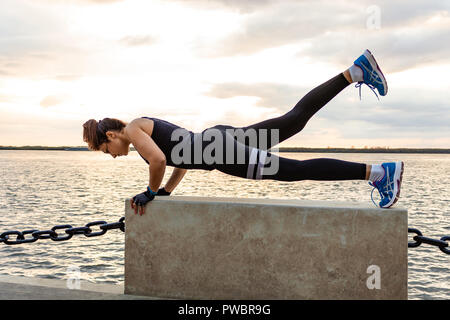 An attractive woman in sports clothes doing sports exercises in nature against the sunset and the Amur river, loves gymnastics, kneads her legs. Active young girl engaged in sports, leads a healthy lifestyle. Stock Photo