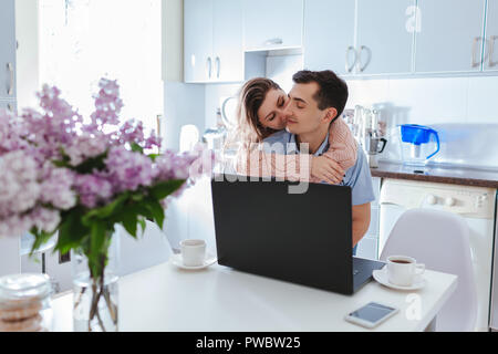 Happy family couple using laptop while having coffee in modern kitchen. Young man and woman hugging and kissing at home Stock Photo