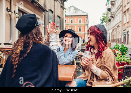 Three female friends having drinks in outdoor cafe. Woman giving high five to her best girlfriend. Women hanging together Stock Photo