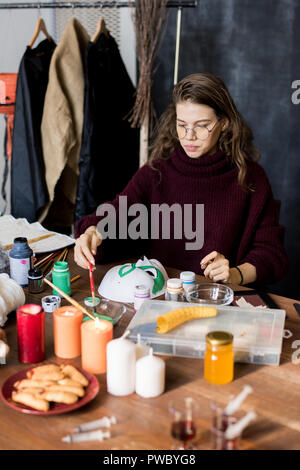 Serious professional female designer in eyeglasses sitting at table and applying paint on brush while working on Halloween mask Stock Photo