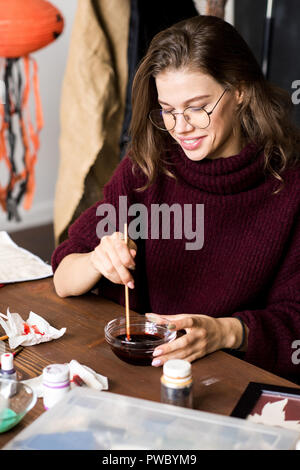 Happy excited creative young female designer in glasses sitting at table and mixing paint to make color of blood for Halloween decoration, she enjoyin Stock Photo