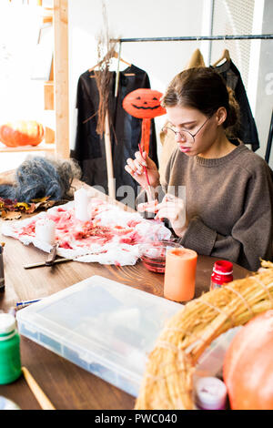 Serious modern young female designer in eyeglasses sitting at table and painting jaw model with red paint while making Halloween costume in workshop Stock Photo