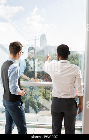 Rear view of young investors in formalwear standing at window and discussing construction project: middle-eastern businessman pointing at building whi Stock Photo