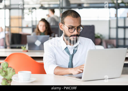 Serious busy handsome middle-eastern manager in eyeglasses sitting at table and using laptop in open space office Stock Photo
