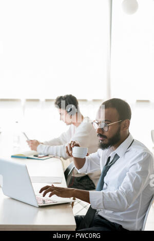 Concentrated busy multi-ethnic managers working in open space office: serious middle-eastern businessman with beard sitting at table and drinking coff Stock Photo