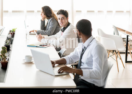 Young people working and resting in coworking space: positive handsome student guy with headphones on neck using tablet and talking to middle-eastern  Stock Photo