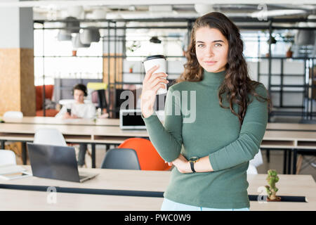 Smiling confident young business lady with curly hair standing at common desk and looking at camera while drinking coffee in open space office Stock Photo