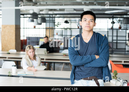 Serious confident handsome Asian guy in jumpsuit standing at table and crossing arms on chest while looking at camera in modern office Stock Photo