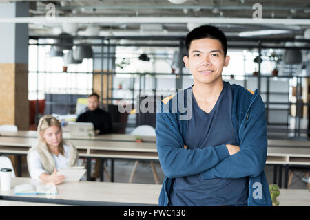 Happy young Asian male intern with crossed arms leaning on table and looking at camera in coworking space Stock Photo