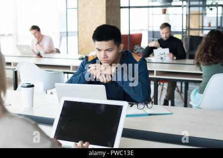 Serious thoughtful Asian male student sitting at table and looking at laptop screen while analyzing data in coworking space Stock Photo