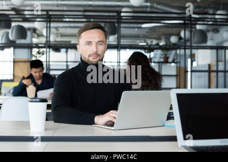Serious confident young bearded businessman in black polo neck sitting at table and looking at camera while working on project in open space office Stock Photo