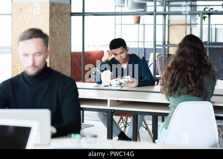 Serious thoughtful young Asian man analyzing papers while reading research and sitting at table with chinese food, people working and eating in open s Stock Photo