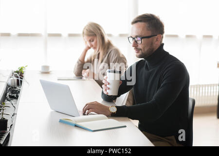 Serious pensive young bearded freelancer in polo neck sitting at table and drinking coffee while using laptop in coworking space Stock Photo
