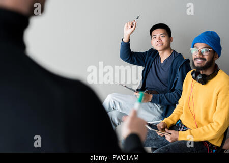 Content curious Asian male student sitting in auditorium and raising hand while having question to speaker at training class Stock Photo