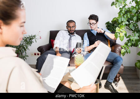 Serious concentrated handsome young multi-ethnic businessmen sitting on sofa in lobby and watching video on smartphone Stock Photo