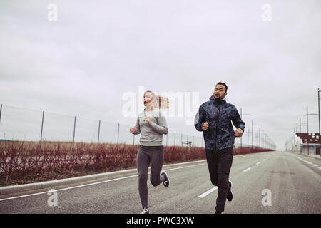 Diverse young couple in sportswear running together along a road in the country on an overcast day Stock Photo