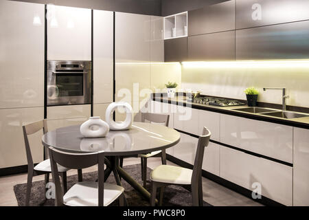 Modern built in kitchen with black and white decor and fitted appliances with circular dining table and chairs set with modern sculptural ceramics Stock Photo